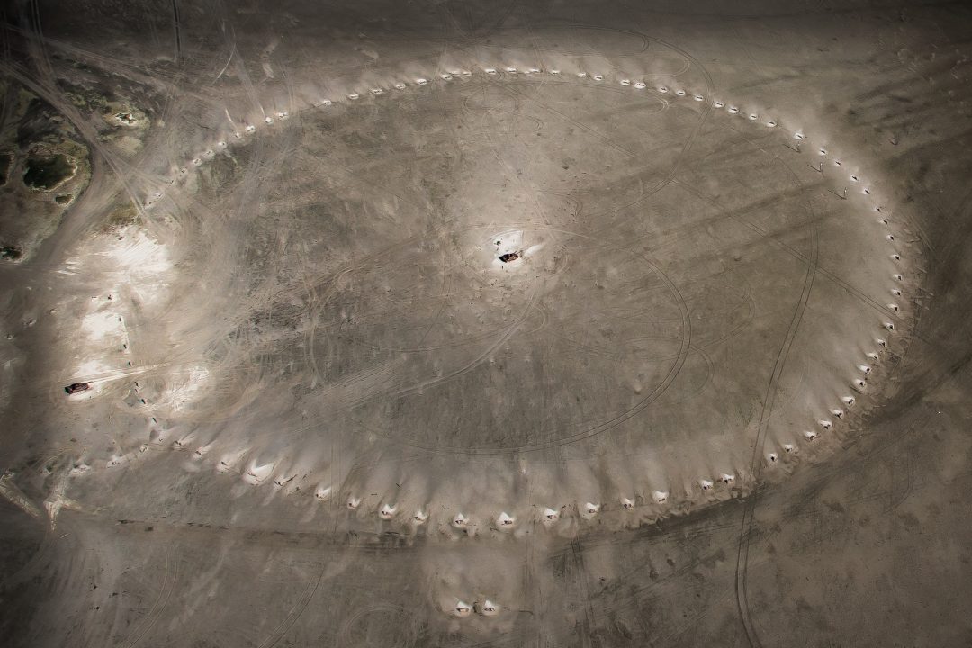 Een cirkel van helmplanten duidelijk zichtbaar vanuit een vliegtuig, door het witte stuifzand. In het midden van de cirkel stank en tank